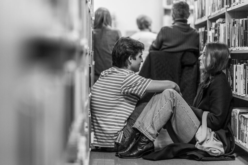 Dwie osoby siedzą na podłodze między regałami w bibliotece i słuchają, a w tle kilka osób siedzą na krzesłach